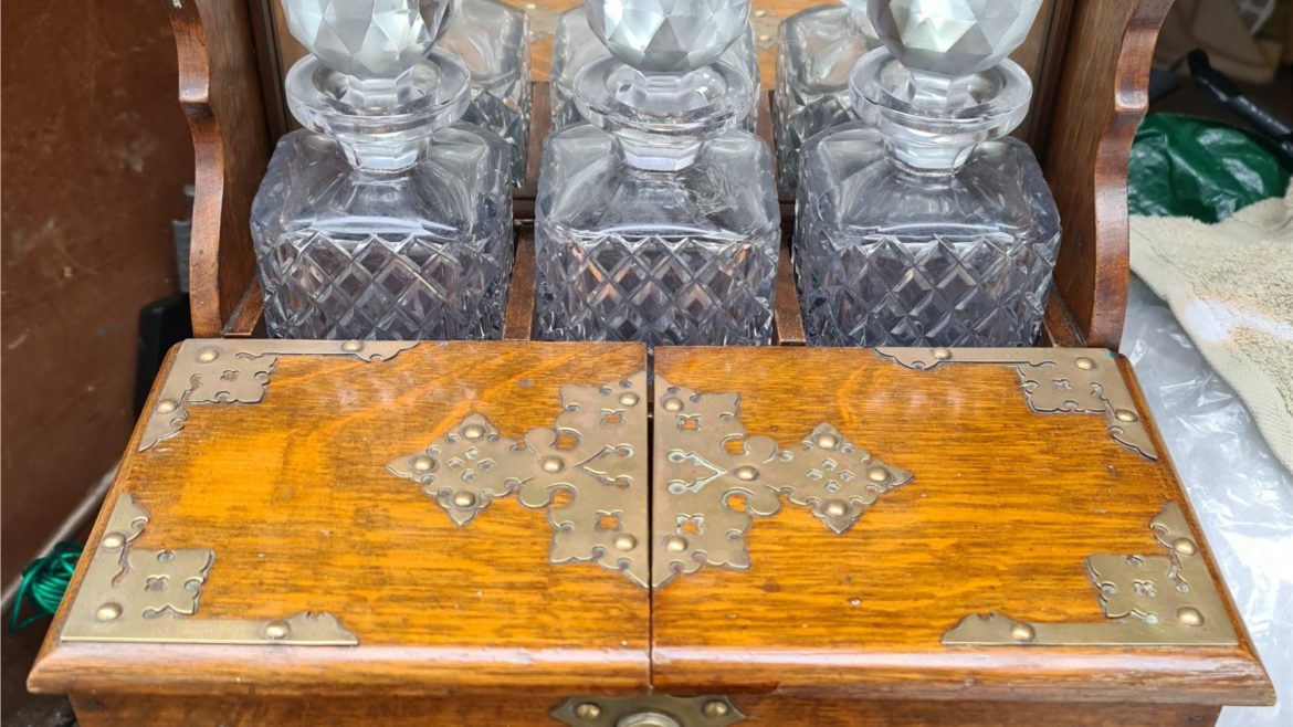 Antique Victorian Games Tantalus With Three Cut Glass Decanters. Oak with brass corner fixings and decorative work. Mirrored back. The front opens up to reveal games pieces. The Tantalus was presented to E. Ainsworth in September 1899 as 1st Prize in the Leamington Spar Bowls Club Handicap. The item measures 35cm by 32cm by 22cm.
