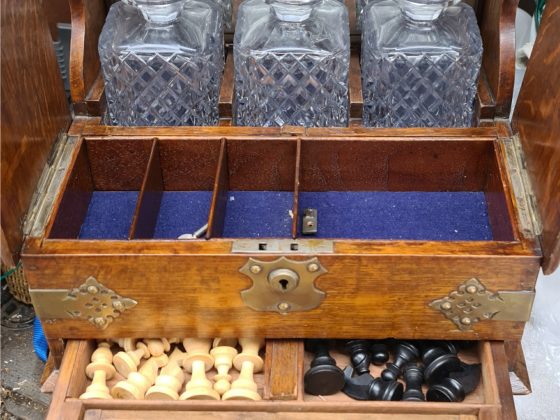 Antique Victorian Games Tantalus With Three Cut Glass Decanters. Oak with brass corner fixings and decorative work. Mirrored back. The front opens up to reveal games pieces. The Tantalus was presented to E. Ainsworth in September 1899 as 1st Prize in the Leamington Spar Bowls Club Handicap. The item measures 35cm by 32cm by 22cm.